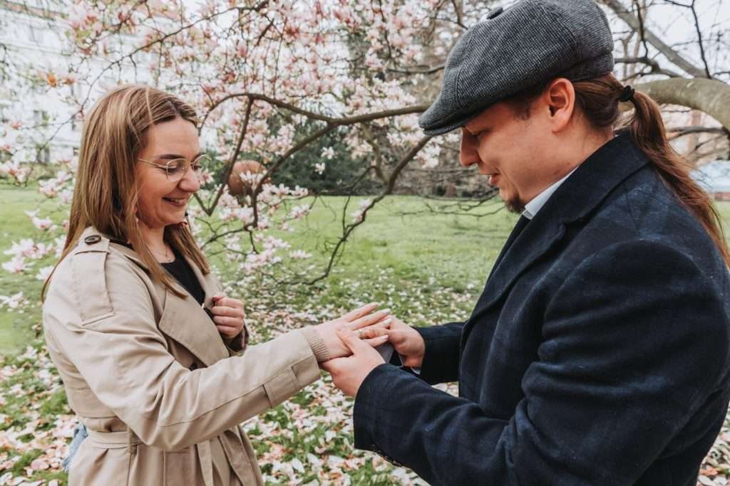 A woman smiling as her partner places an engagement ring on her finger under blossoming trees
