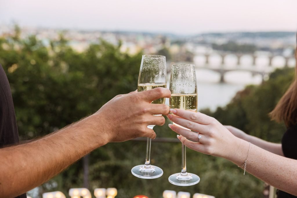 Two champagne glasses raised in a toast after a proposal, with an engagement ring visible and Prague's bridges in the background